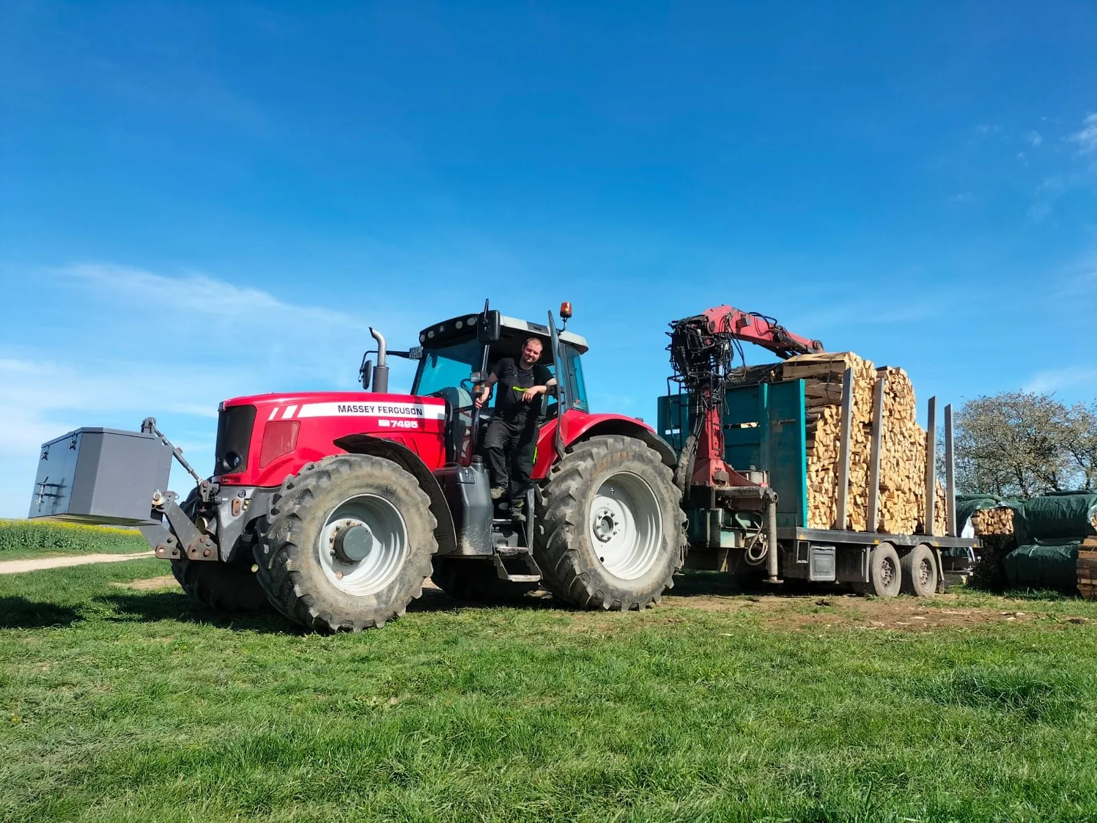 Mann in Arbeitskleidung am Aufstieg des Massey Ferguson 7495, dahinter der mit Scheitholz beladene Rückewagen